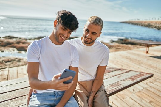 Young gay couple smiling happy using smartphone at the beach promenade.
