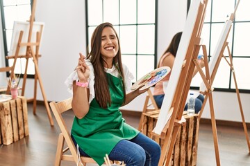 Young hispanic artist women painting on canvas at art studio smiling and laughing with hand on face covering eyes for surprise. blind concept.
