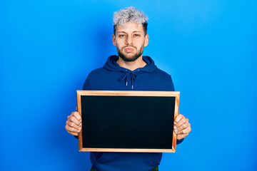 Young hispanic man with modern dyed hair holding blackboard puffing cheeks with funny face. mouth...