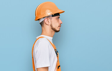 Hispanic young man wearing handyman uniform and safety hardhat looking to side, relax profile pose with natural face with confident smile.