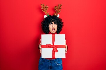 Young middle east woman wearing deer christmas hat holding gift angry and mad screaming frustrated and furious, shouting with anger. rage and aggressive concept.