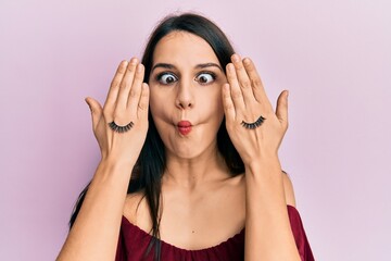 Fototapeta premium Young hispanic woman covering eyes with hands and fake lashes making fish face with mouth and squinting eyes, crazy and comical.