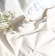 Organic cotton white bed sheets concept frame with chamomile flowers and a sleeping mask. Copy space