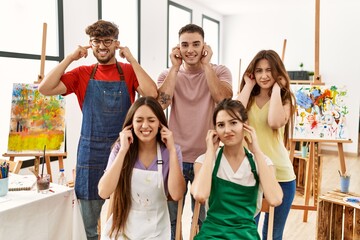 Group of five hispanic artists at art studio covering ears with fingers with annoyed expression for the noise of loud music. deaf concept.