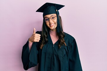Young hispanic woman wearing graduation cap and ceremony robe doing happy thumbs up gesture with hand. approving expression looking at the camera showing success.