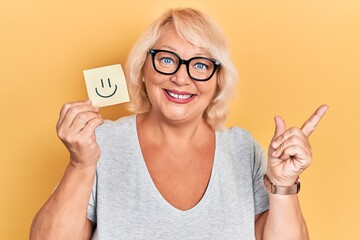 Middle age blonde woman holding smile reminder smiling happy pointing with hand and finger to the side