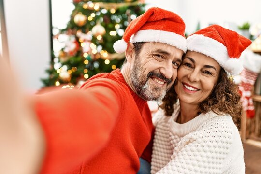 Middle age hispanic couple smiling happy wearing christmas hat. Sitting on the floor making selfie by the camera at home.