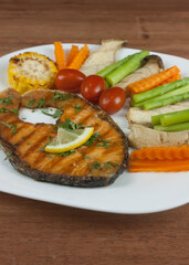 Grilled salmon steak slides until cooked, placed on a white plate with lemon on fish and vegetables, placed around the dish on a wooden floor.
