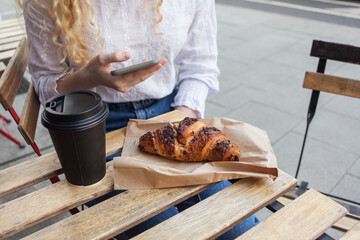 Young white woman having a break, veranda table in cafeteria with cardboard cup of coffee to go and french croissant, holds smartphone in hand. quick snack, business city street breakfast or brunch