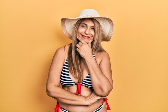 Middle age hispanic woman wearing bikini and summer hat looking confident at the camera smiling with crossed arms and hand raised on chin. thinking positive.
