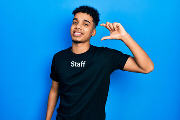 Young african american man wearing staff t shirt smiling and confident gesturing with hand doing small size sign with fingers looking and the camera. measure concept.