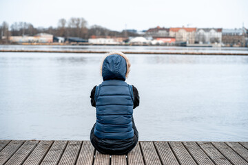 Young girl in black jeans and a blue warm vest with a hood sits with her back on a wooden pier by...