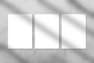 Three empty white vertical rectangle poster or business card mockups with window shadow on the gray concrete stone wall. Flat lay, top view. For advertising, brand design, stationery presentation.