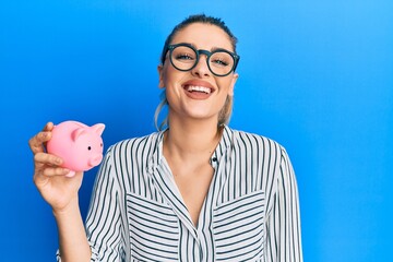 Young caucasian woman wearing business clothes holding piggy bank looking positive and happy standing and smiling with a confident smile showing teeth