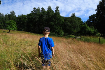 Naklejka na ściany i meble Boy with blue shirt and cap standing in the middle of a field during the summer. Sunny day outside. Great vista or meadow. Green trees in the background. Stockholm, Sweden, Europe.