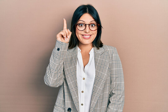 Young hispanic woman wearing business clothes and glasses pointing finger up with successful idea. exited and happy. number one.
