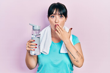 Young hispanic woman wearing sportswear holding water bottle covering mouth with hand, shocked and afraid for mistake. surprised expression