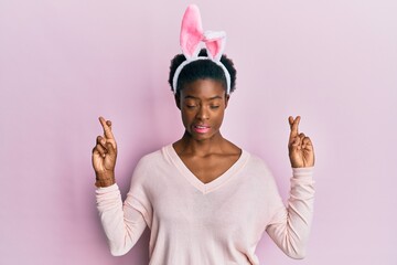 Young african american girl wearing cute easter bunny ears gesturing finger crossed smiling with hope and eyes closed. luck and superstitious concept.