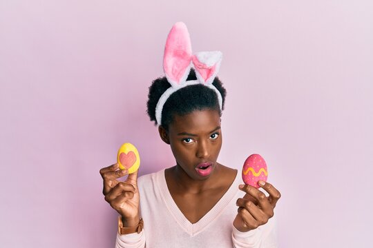 Young african american girl wearing cute easter bunny ears holding painted eggs in shock face, looking skeptical and sarcastic, surprised with open mouth