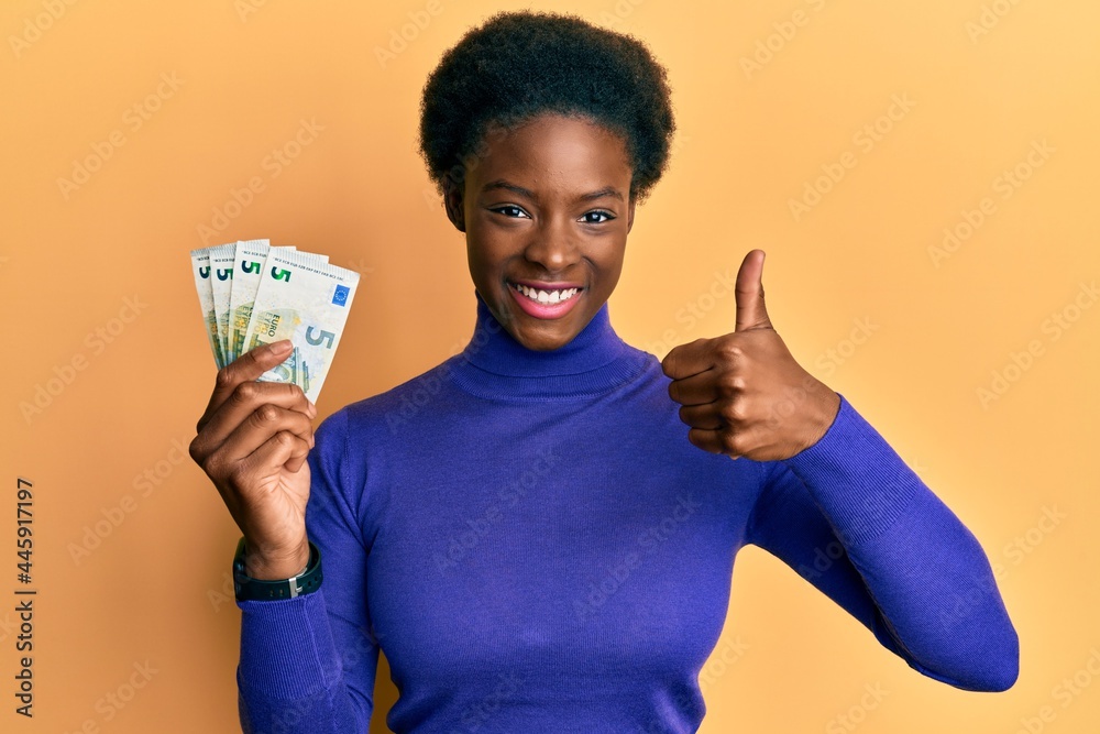 Poster Young african american girl holding bunch of 5 euro banknotes smiling happy and positive, thumb up doing excellent and approval sign