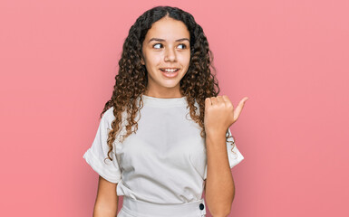 Teenager hispanic girl wearing casual white t shirt smiling with happy face looking and pointing to the side with thumb up.