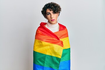 Young man wearing woman make up wrapped in rainbow lgbtq flag relaxed with serious expression on face. simple and natural looking at the camera. - obrazy, fototapety, plakaty