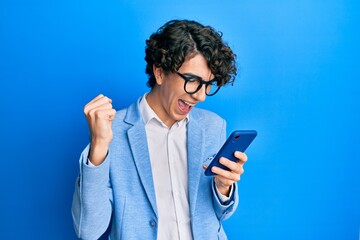 Fototapeta premium Hispanic young man using smartphone wearing business jacket screaming proud, celebrating victory and success very excited with raised arms