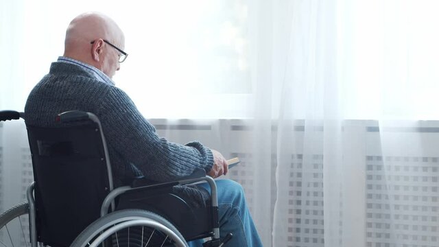 Disabled old man is sitting in a wheelchair at home alone. A handicapped person in a wheelchair is reading the book. Paralysis and disability.