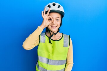 Beautiful brunette little girl wearing bike helmet and reflective vest doing ok gesture with hand smiling, eye looking through fingers with happy face.