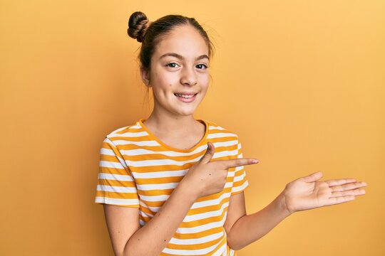 Beautiful brunette little girl wearing casual striped t shirt amazed and smiling to the camera while presenting with hand and pointing with finger.