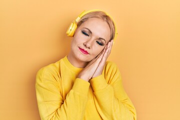 Young blonde woman listening to music using headphones sleeping tired dreaming and posing with hands together while smiling with closed eyes.