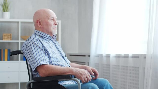 Disabled old man is sitting in a wheelchair at home alone. A handicapped person in a wheelchair is looking out the window. Paralysis and disability.