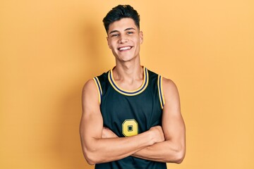 Young hispanic man wearing basketball uniform happy face smiling with crossed arms looking at the...