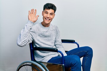Young hispanic man sitting on wheelchair waiving saying hello happy and smiling, friendly welcome...