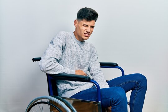 Young hispanic man sitting on wheelchair with hand on stomach because indigestion, painful illness feeling unwell. ache concept.