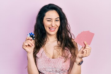 Young hispanic woman playing poker holding casino chips and cards winking looking at the camera...