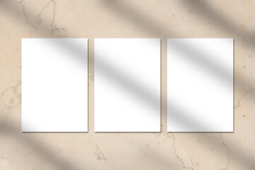 Three empty white vertical A4 rectangle poster or business card mockups with window shadow on beige marble stone wall. Flat lay, top view. For advertising, brand design, stationery presentation.