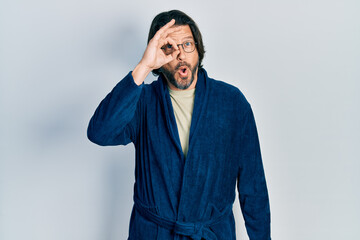 Middle age caucasian man wearing bathrobe and glasses doing ok gesture shocked with surprised face, eye looking through fingers. unbelieving expression.