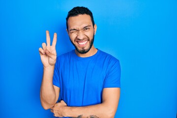 Naklejka premium Hispanic man with beard wearing casual blue t shirt smiling with happy face winking at the camera doing victory sign. number two.