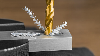Closeup of spiral fluted drill bit and screw shavings in metal part on brown background. Drilling hole in aluminum profile clamped in black jaws of bench vise detail. Sharp titanium coated steel tool.