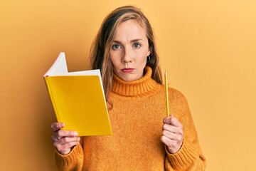Young blonde woman holding book and pencil skeptic and nervous, frowning upset because of problem....