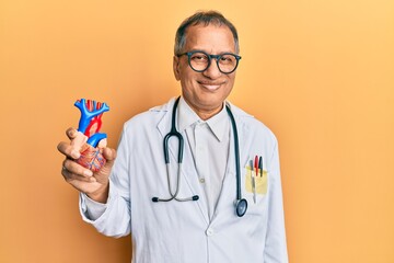 Middle age indian man wearing doctor uniform holding heart looking positive and happy standing and smiling with a confident smile showing teeth
