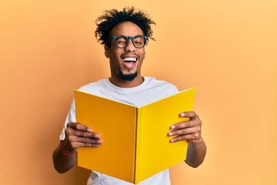 Young african american man with beard reading a book wearing glasses winking looking at the camera with sexy expression, cheerful and happy face.