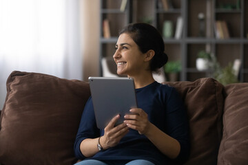 Happy millennial Indian woman relax on couch at home use modern pad gadget look in distance dreaming thinking. Smiling young biracial female client hold tablet talk on video webcam call on device.