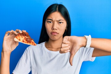 Young chinese woman eating tasty pepperoni pizza with angry face, negative sign showing dislike with thumbs down, rejection concept