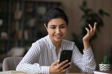 Smiling young Indian woman in headphones talk speak on video call on cellphone gadget. Happy mixed...