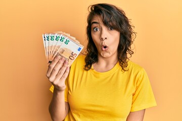 Young hispanic woman holding bunch of 50 euro banknotes scared and amazed with open mouth for...