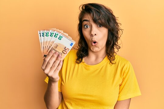 Young hispanic woman holding bunch of 50 euro banknotes scared and amazed with open mouth for surprise, disbelief face