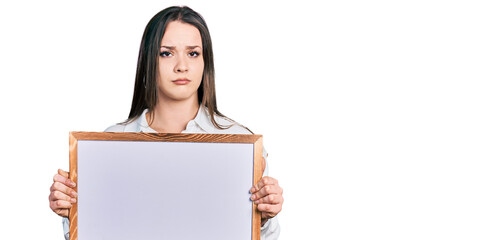 Young hispanic woman holding empty white chalkboard clueless and confused expression. doubt concept.