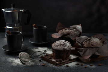 Chocolate coffee muffins with powdered sugar and cinnamon and cups with a drink on a dark background. Low key